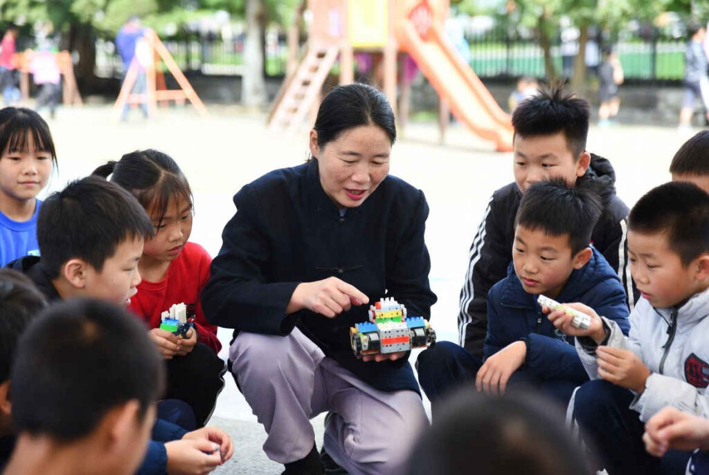 Female village teacher coaching children to assemble a robot in a playground